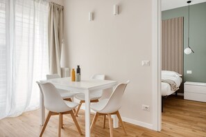 A modern, well-lit room with a white dining table, four chairs, and a bed with a striped headboard. - Hostmate - Milan - Imperia's Serenity - Romolo (Milan)