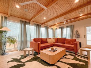 A cozy living room with a wooden ceiling, a sectional sofa, a coffee table, and a large window with curtains. - Harlow’s Hideaway – Cozy Retro Flat-Top Near Beach (Kill Devil Hills)
