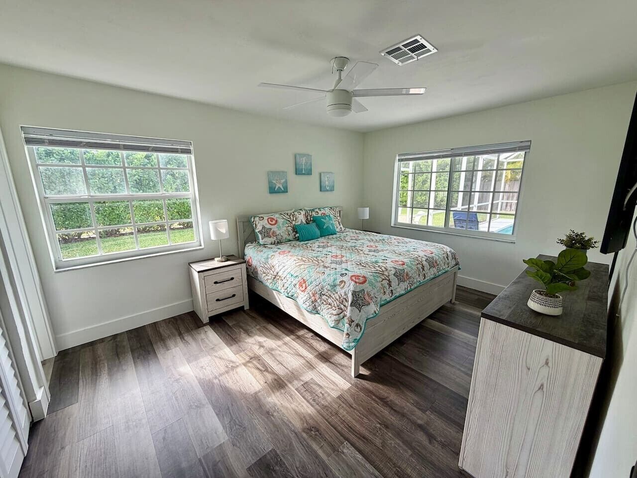 A bedroom with a bed, two bedside tables, a nightstand, a ceiling fan, and two windows.