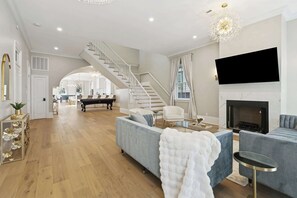 A spacious living room with a grand staircase, a fireplace, and a flat-screen TV mounted on the wall. - Luxury Estate w/ Private Pool, Spa & Gated Parking (New Orleans)