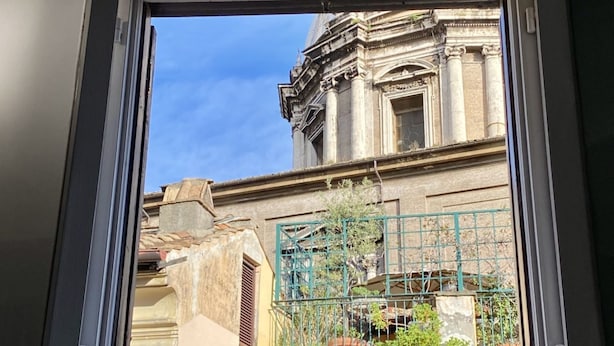 Blick durch ein Fenster auf ein Gebäude mit klassischer Architektur, darunter Säulen und ein Giebel, sowie einen grünen Metallzaun mit Blumentöpfen.