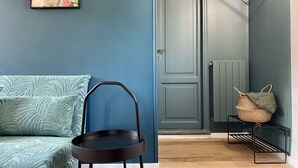 Ein Zimmer mit einer türkisfarbenen Wand, einem Bett mit gemusterten Textilien, einem schwarzen Metallstuhl, einer Tür und einem Heizkörper.