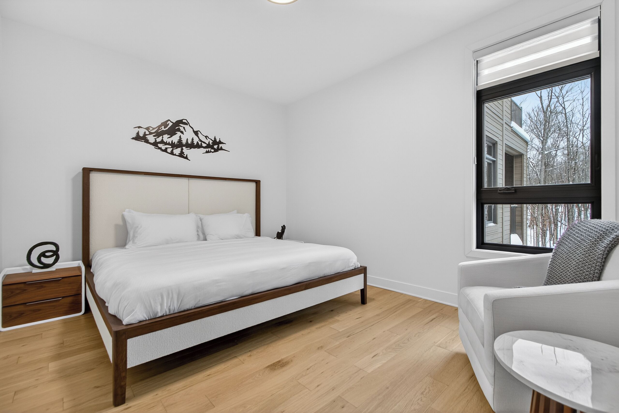 A bedroom with a wooden bed, a nightstand, a chair, and a window with a view of buildings and trees.