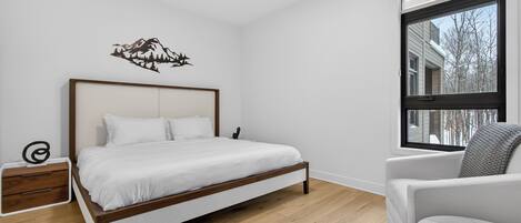 A bedroom with a wooden bed, a nightstand, a chair, and a window with a view of buildings and trees.
