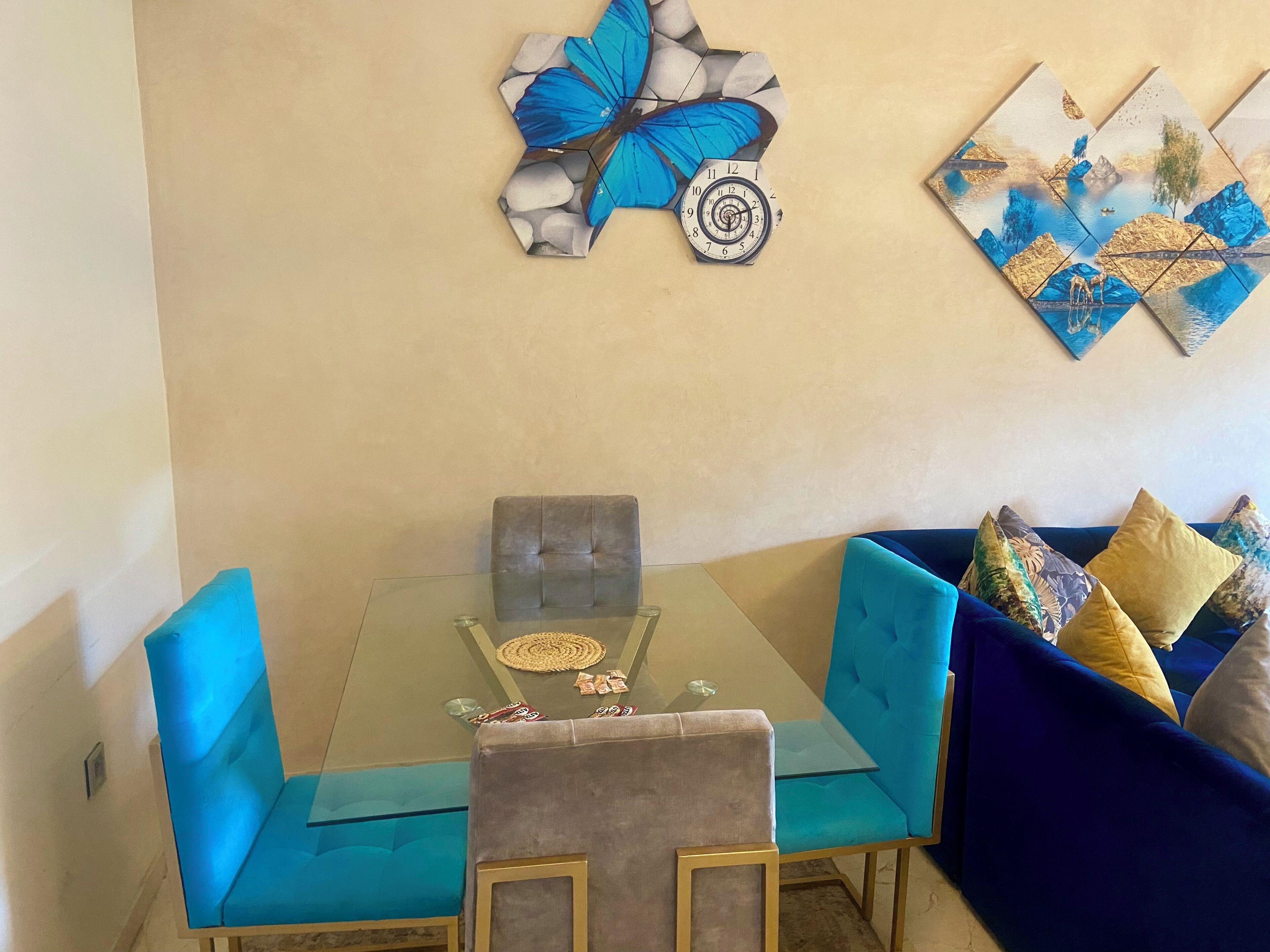 A dining area with a glass table, two blue upholstered chairs, a grey upholstered chair, and a blue sofa with patterned cushions.