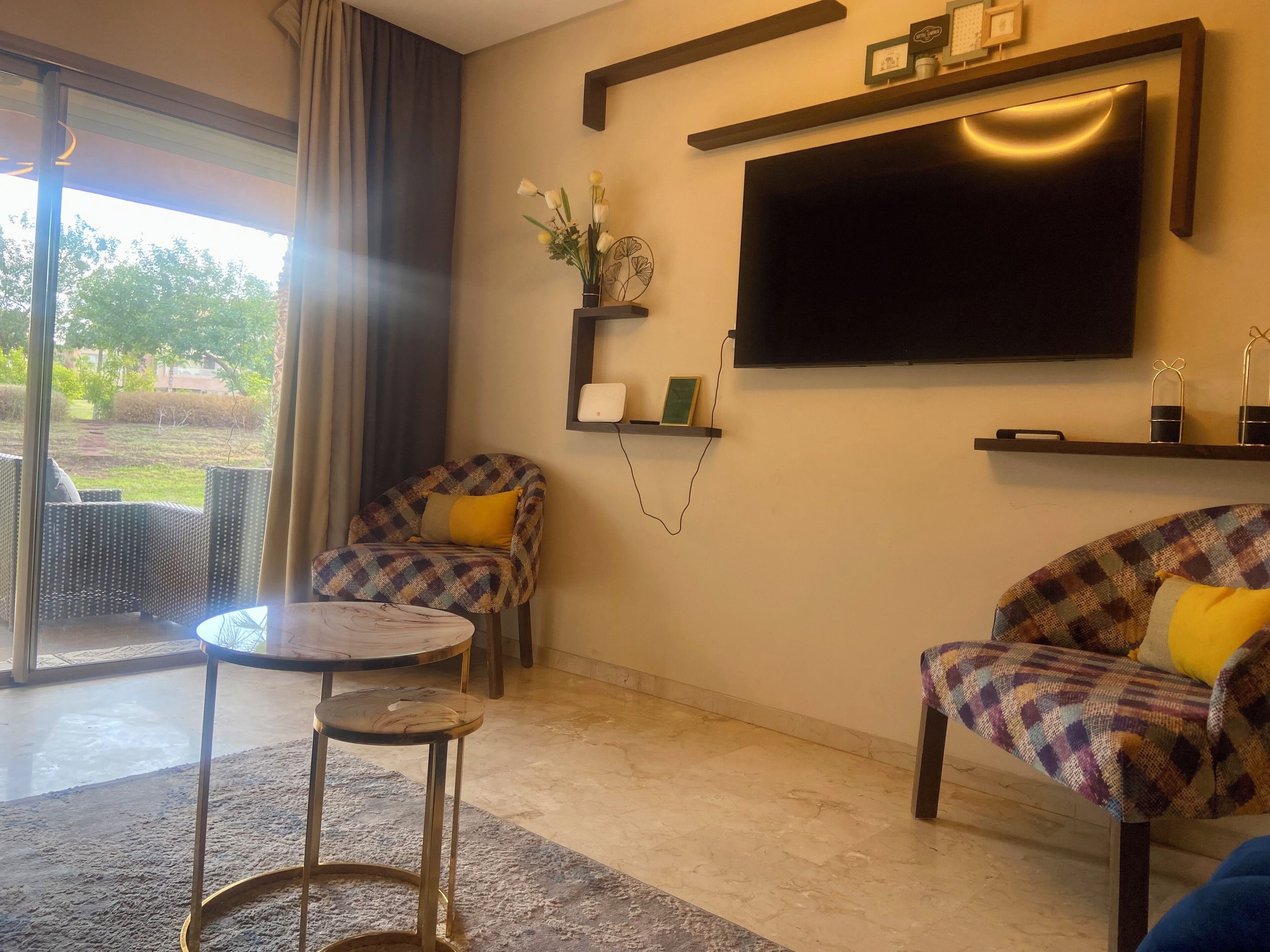 A living room with a sofa, armchair, and a small table. There is a TV mounted on the wall and a window with curtains.