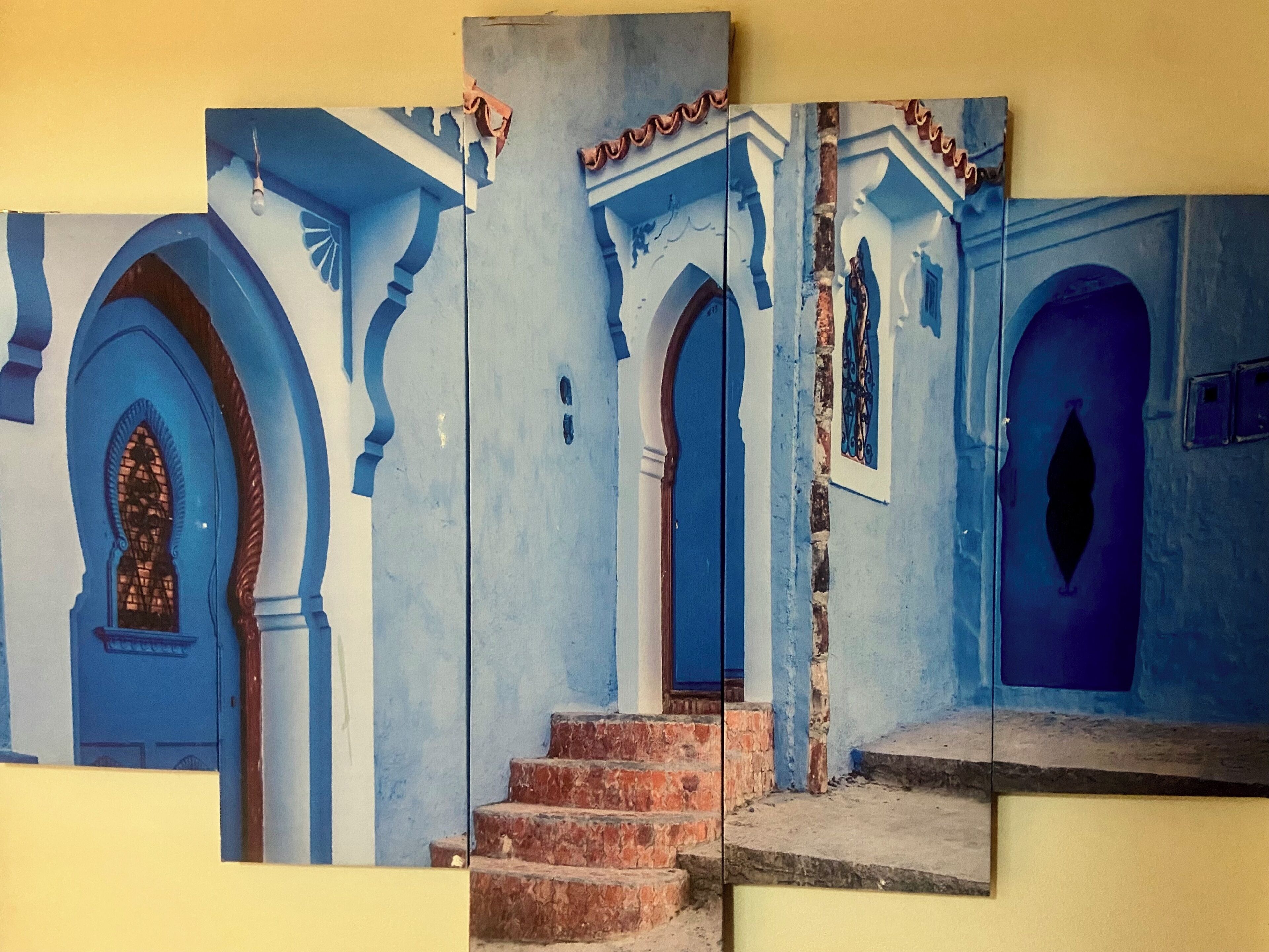 Obra de arte compuesta por varios paneles que muestra la fachada de un edificio con puertas y arcos azules, una ventana con celosía decorativa y una escalera que conduce a una puerta.