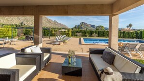 Un patio exterior con mobiliario de mimbre, una piscina y montañas al fondo.