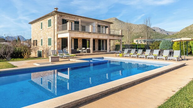 Una casa de piedra de dos pisos con piscina, mobiliario exterior y montañas al fondo.