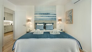 Ein modernes Hotelzimmer mit einem großen Bett, zwei Nachttischlampen, einem Gemälde an der Wand und Blick auf den Küchenbereich.