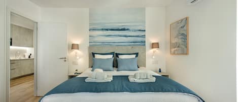 A modern hotel room with a large bed, two bedside lamps, a painting on the wall, and a view of the kitchen area.