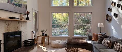 A living room with a fireplace, a sofa, a coffee table, and large windows with a view of trees.