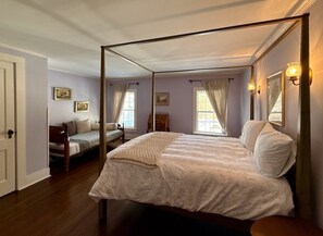 A four-poster bed with a white bedspread, a wooden floor, a chair with a cushion, and a small table.