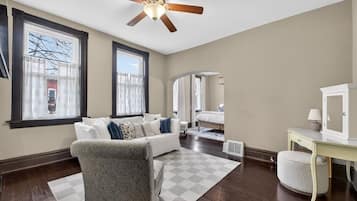 A spacious living room with a white sofa, patterned armchair, and a checkered rug. A ceiling fan and a white cabinet are visible.