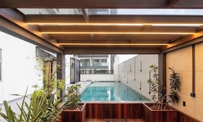 Un espace piscine intérieure moderne, doté d’une terrasse en bois, de plantes en pot et offrant une vue sur un bâtiment en arrière-plan.