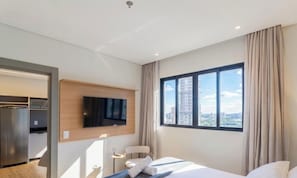 Une chambre d’hôtel moderne équipée d’un lit, d’un téléviseur fixé au mur, d’une fenêtre donnant sur la ville et d’une kitchenette avec un réfrigérateur et un four à micro-ondes.
