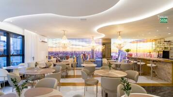 A modern hotel interior with round tables, chairs, chandeliers, a cityscape mural, and a bar area.