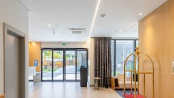 A hotel lobby with a reception desk, a seating area with a sofa and armchairs, a large window with a view of the outdoors, and a red carpet leading to the entrance.