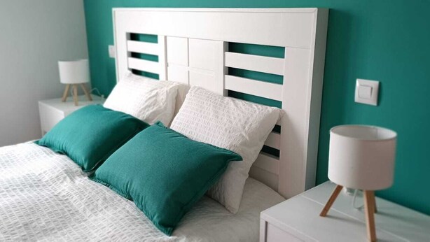 A neatly made bed with white and teal bedding, a white headboard with horizontal slats, and a bedside table with a white lamp.