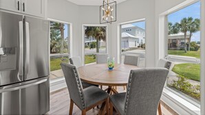 A modern kitchen with a stainless steel refrigerator, a wooden dining table surrounded by chairs, and large windows offering a view of the neighborhood. - Blue Mountain Home, Pet friendly, Pool, 4 Bikes, Beach (Santa Rosa Beach)