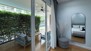 Une chambre d’hôtel moderne dotée d’une porte vitrée donnant sur un balcon, d’un pouf rond, d’un grand miroir et d’un lit agrémenté d’un coussin décoratif noir.