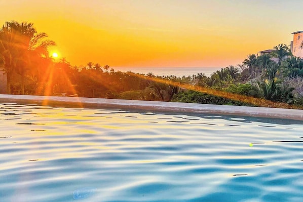 Une piscine offrant une vue sur le coucher du soleil, entourée de palmiers et avec un bâtiment en arrière-plan.