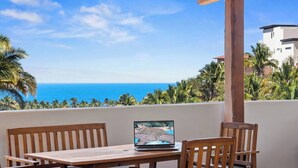 Una mesa de madera con un ordenador portátil, con vista a la costa, palmeras y un cielo azul despejado.