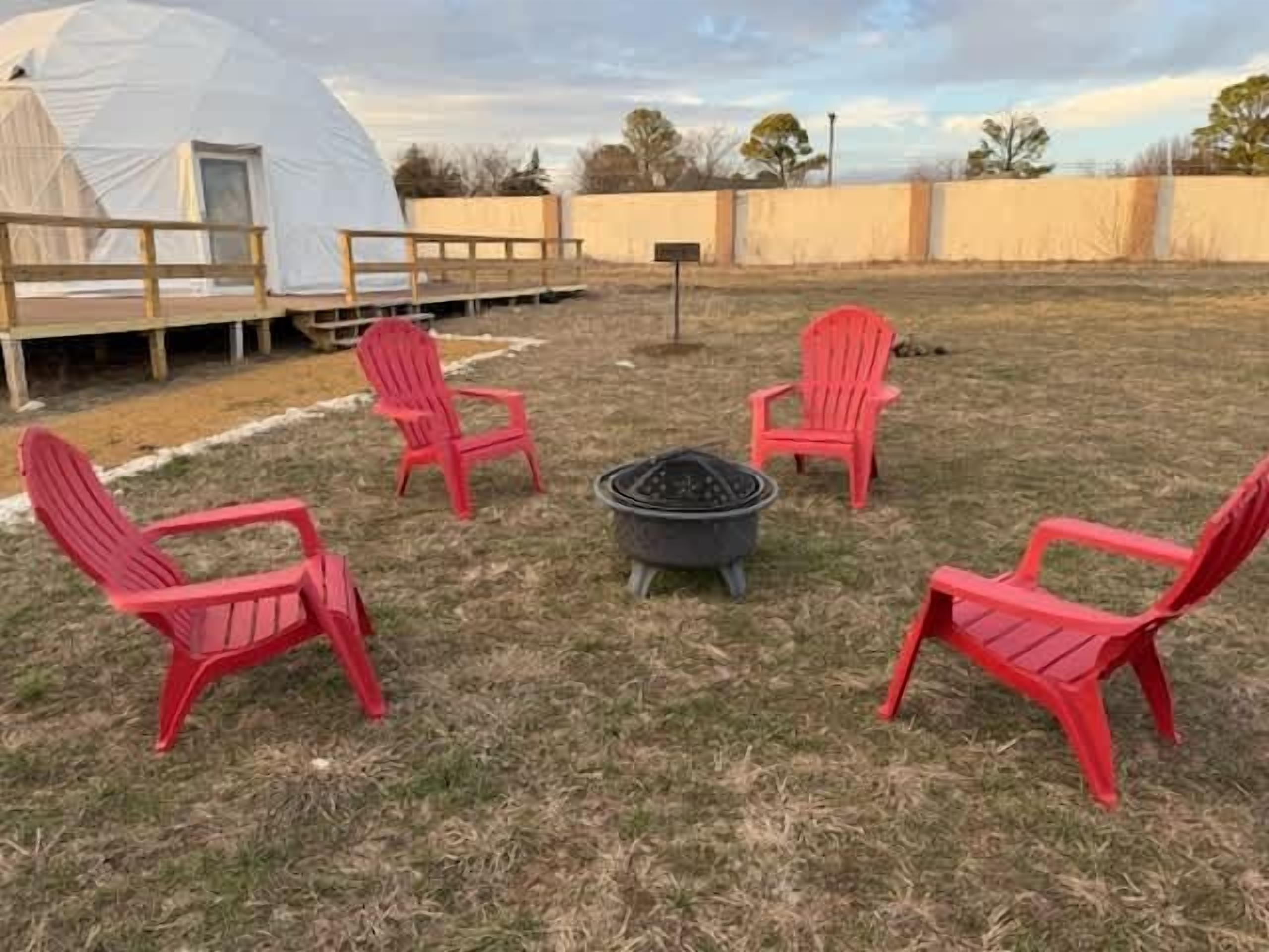 A tented outdoor area with four red chairs arranged around a central fire pit.