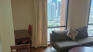 Una habitación con una mesa de comedor de madera, un sofá con un cojín estampado, una pequeña mesa redonda y vistas a edificios altos a través de una ventana con cortinas.