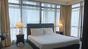 Habitación de hotel con una cama grande, dos lámparas de noche, una mesita y vistas a la ciudad a través de amplios ventanales.