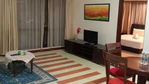 Habitación de hotel con un ventanal grande, un televisor de pantalla plana sobre un pedestal negro, una mesa redonda pequeña con mantel floral, una silla de madera con cojines rojos, una alfombra azul estampada y un cuadro en la pared.