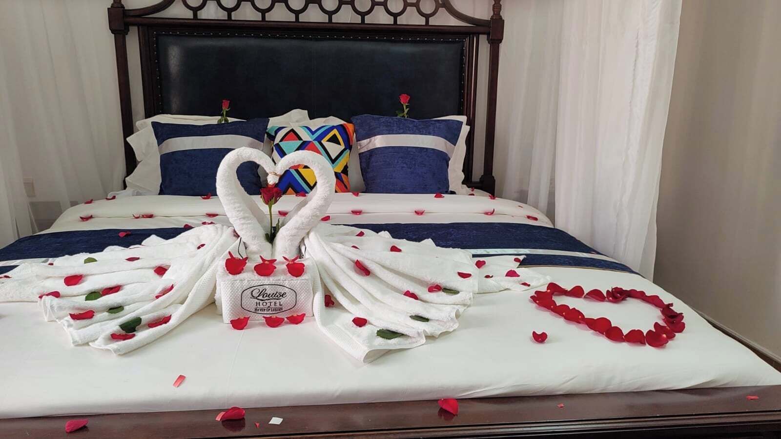 Una cama bien arreglada con una toalla dispuesta en forma de corazón, un ramo de rosas rojas y una decoración floral con forma de corazón sobre la cama.