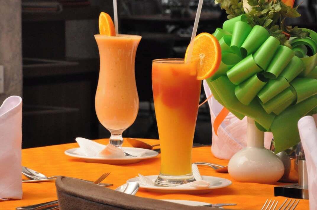 Une table dressée avec deux grands verres de jus d’orange, un vase blanc contenant des feuilles vertes et un porte-serviettes marron.