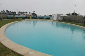 A large, oval-shaped swimming pool with a concrete edge, surrounded by a grassy area and a wooden fence.