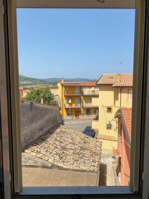 Vista des d'una finestra amb una perspectiva d'un carrer amb edificis, cel blau clar i muntanyes allunyades.