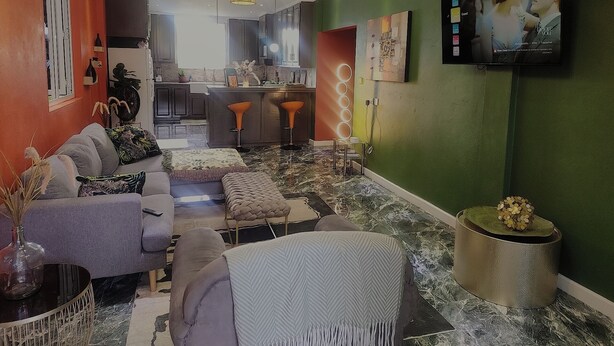 Ein modernes Wohnzimmer mit einer Couch, einem Hocker und einem Couchtisch. Im Hintergrund ist eine Küche mit Essbereich zu sehen.