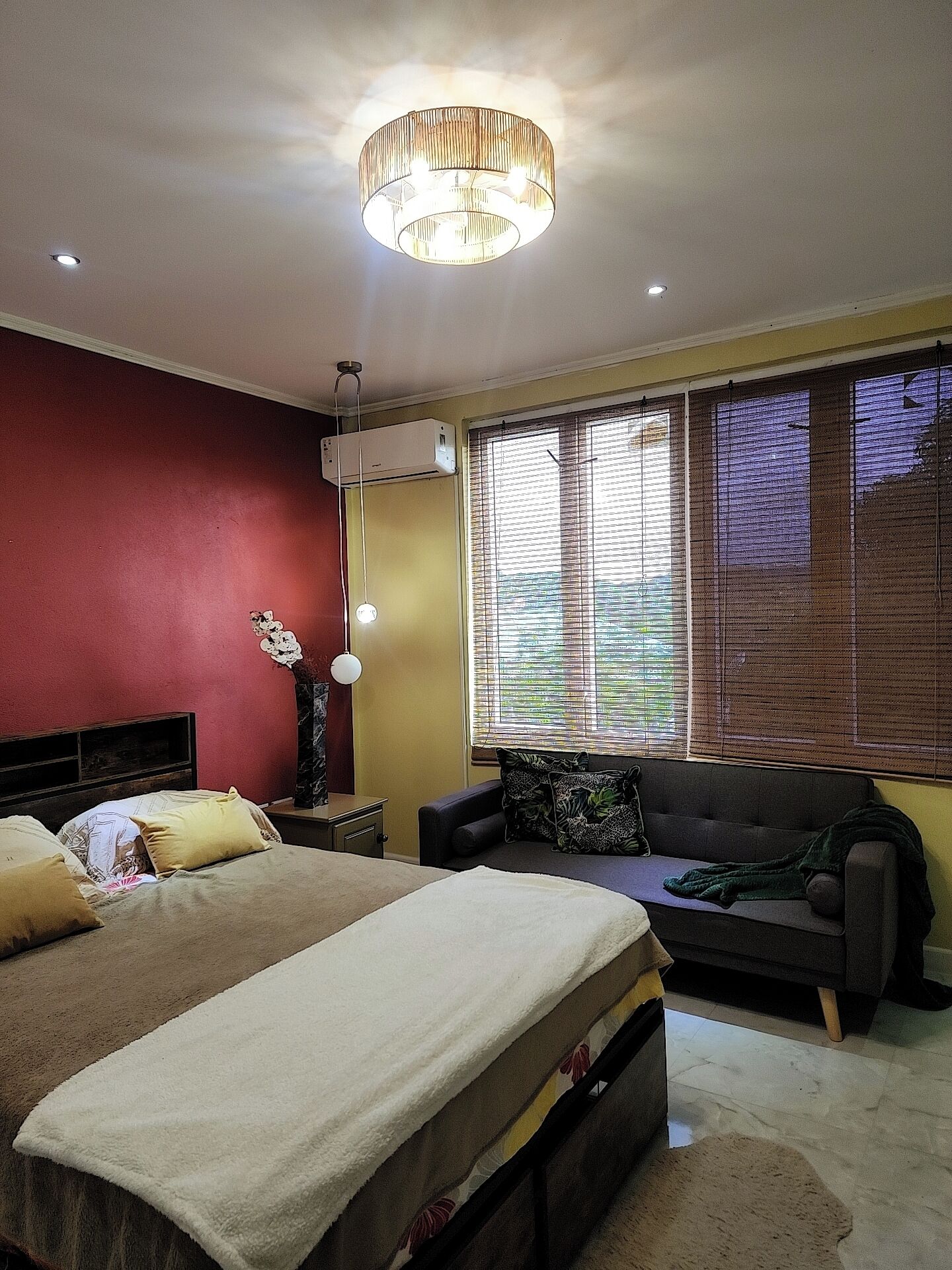 A bedroom with a bed, a sofa, a ceiling light, and a window with blinds.