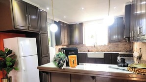 Eine moderne Küche mit dunklen Schränken, einem weißen Kühlschrank, einer Spüle und einem Herd. Zwei orangefarbene Barhocker und ein Blumentopf sind ebenfalls vorhanden.
