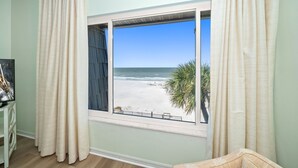 Ein Zimmer mit großem Fenster mit Blick auf einen Strand, Vorhänge an beiden Seiten und ein kleiner Tisch mit einem Bilderrahmen.