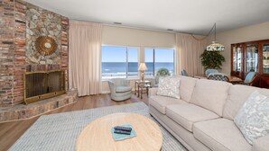 Ein Wohnzimmer mit Kamin aus Stein, einem beigen Sofa, einem runden Couchtisch und Meerblick durch große Fenster.