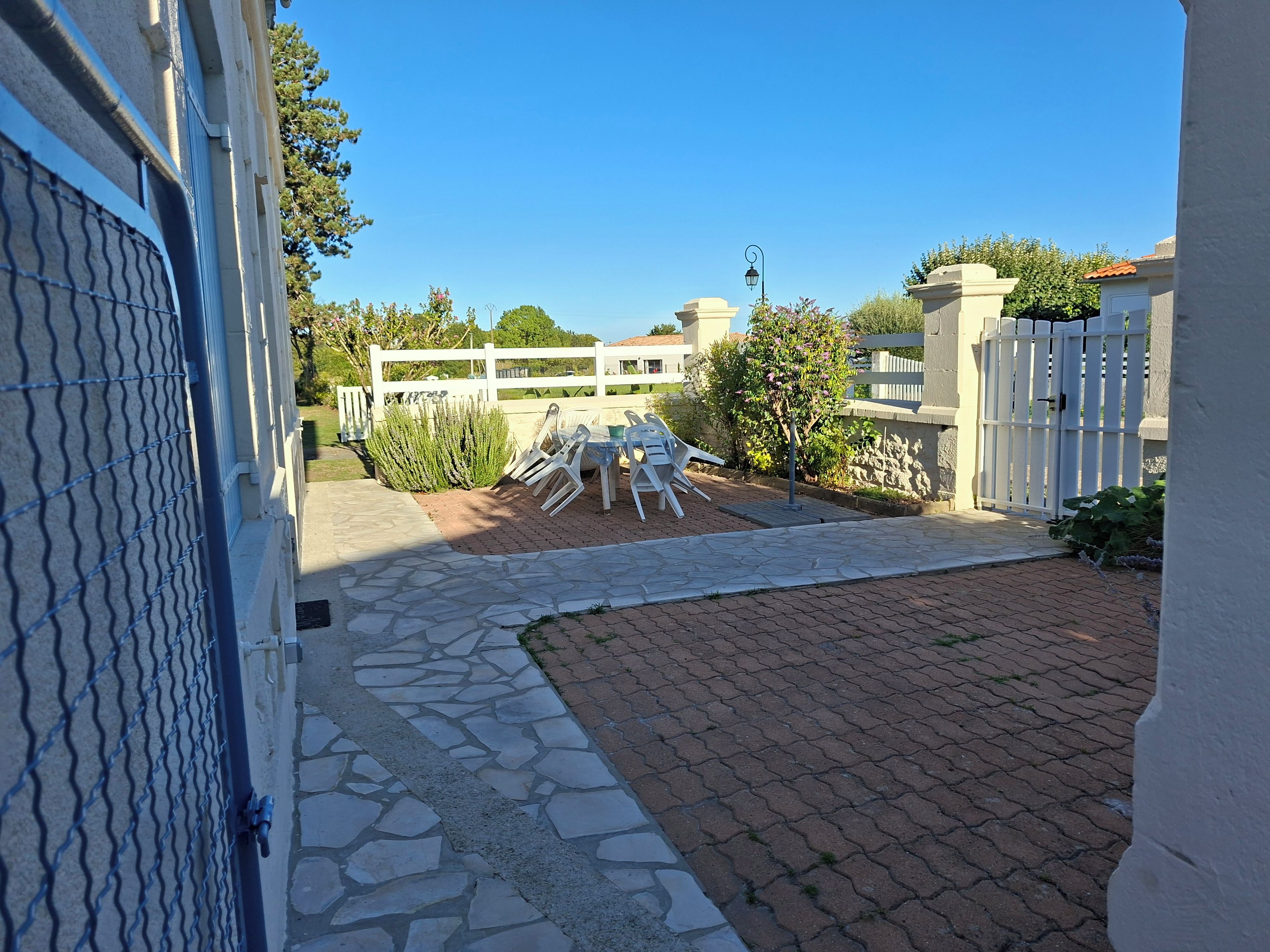 A paved patio area with a white fence, a white gate, and a row of white plastic chairs.