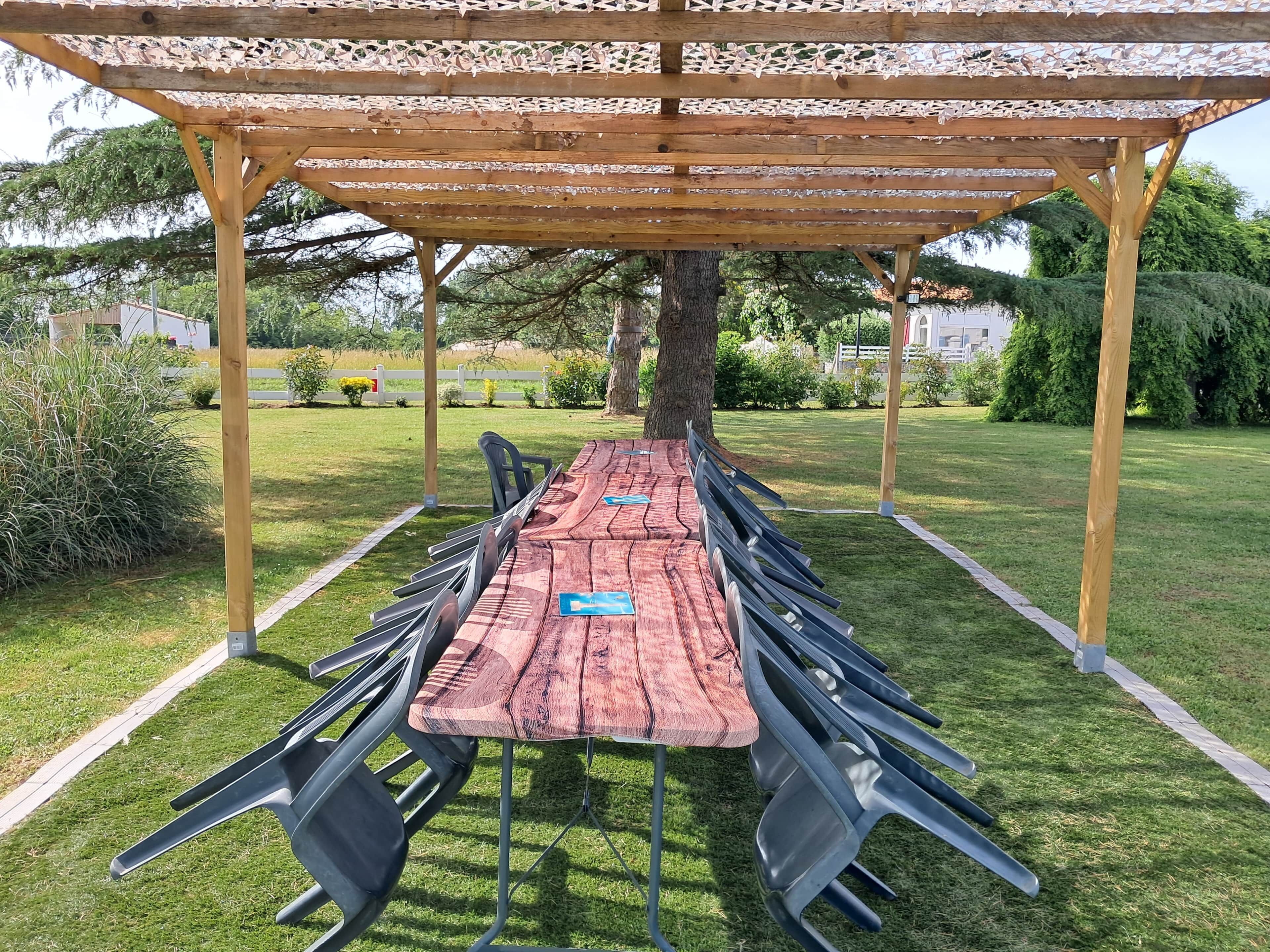 Un espace extérieur abrité, aménagé pour les repas, avec une longue table et des chaises, entouré d’une pelouse verdoyante et d’arbres.