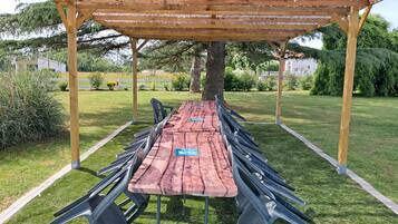 Un espace extérieur abrité, aménagé pour les repas, avec une longue table et des chaises, entouré d’une pelouse verdoyante et d’arbres.