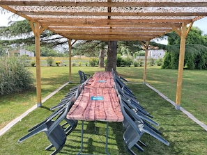 Un espace extérieur abrité, aménagé pour les repas, avec une longue table et des chaises, entouré d’une pelouse verdoyante et d’arbres.