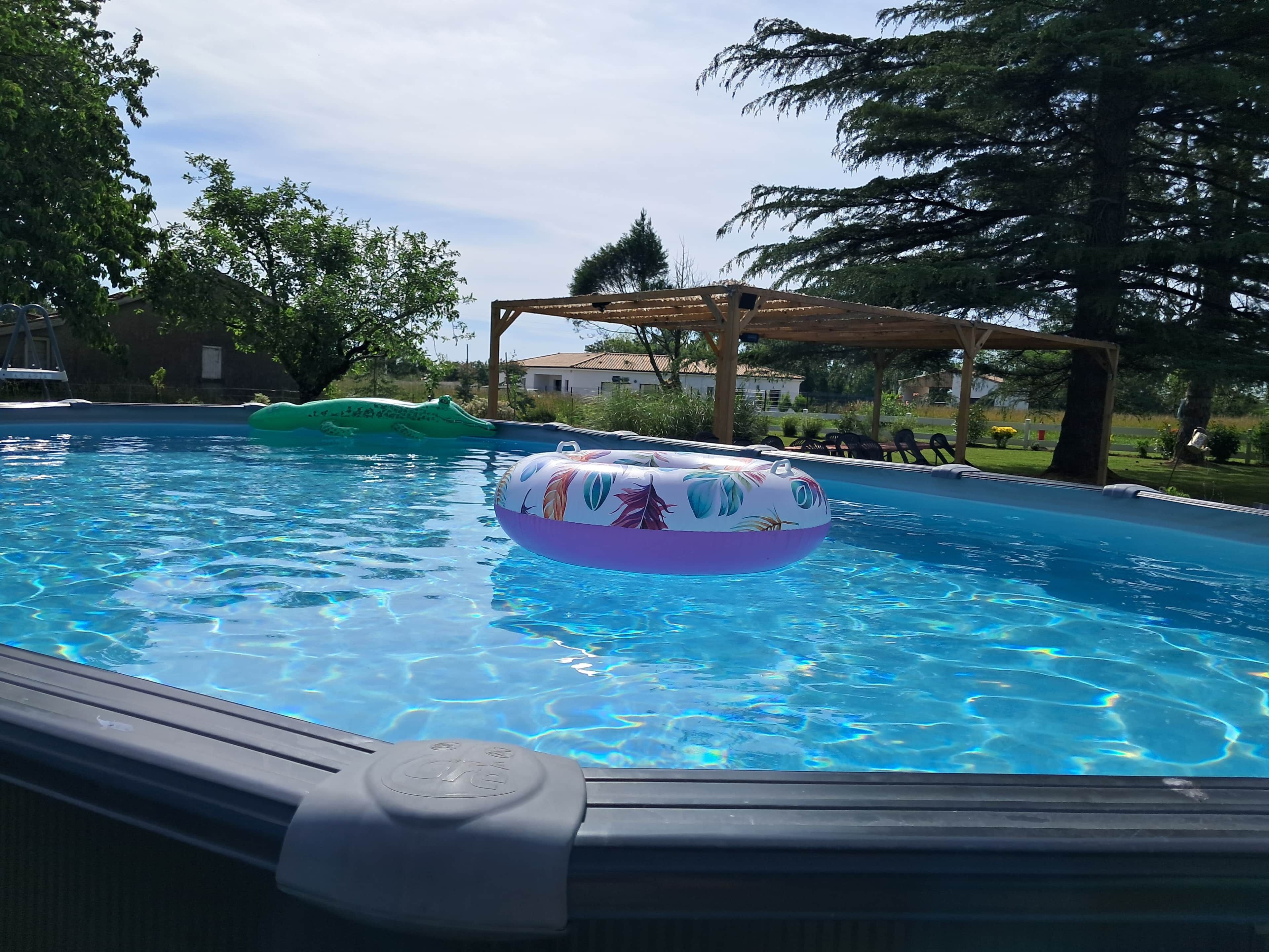 Une piscine avec des jeux gonflables, entourée d’arbres et agrémentée d’une pergola en bois.