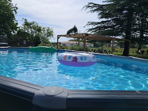 Une piscine avec des jeux gonflables, entourée d’arbres et agrémentée d’une pergola en bois.