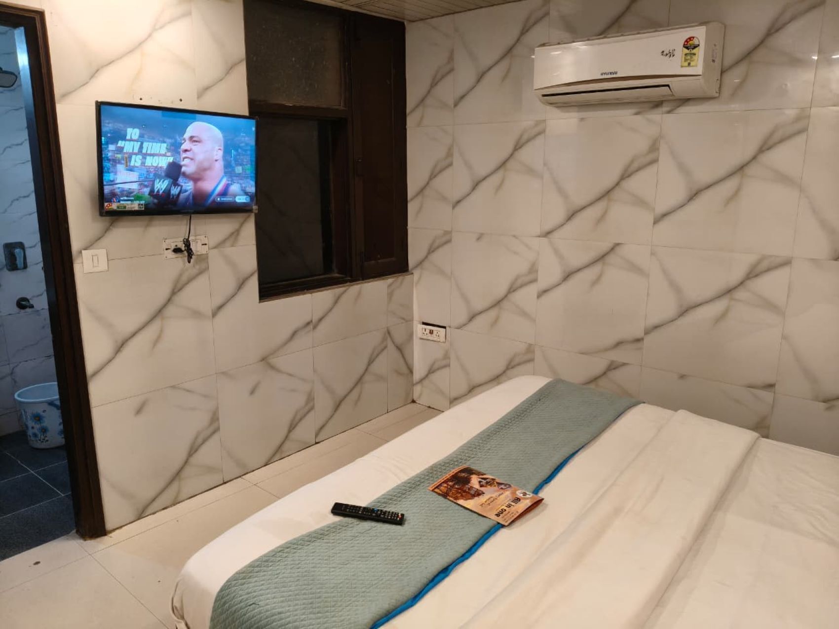 A hotel room with a bed, a TV mounted on the wall, and an air conditioning unit.