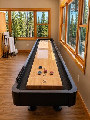 Una mesa de shuffleboard con tres discos en juego, frente a grandes ventanales con vistas a un bosque.