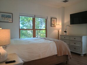 Ein Schlafzimmer mit Bett, Nachttischlampe, Schminktisch, Fernseher und einem Fenster mit Blick auf Bäume.