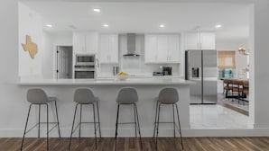 A modern kitchen with white cabinets, a large island with bar stools, and a dining area with a wooden table and chairs. - Backyard Oasis: Pool, Hot Tub, Giant Chess & Golf (Fort Worth)